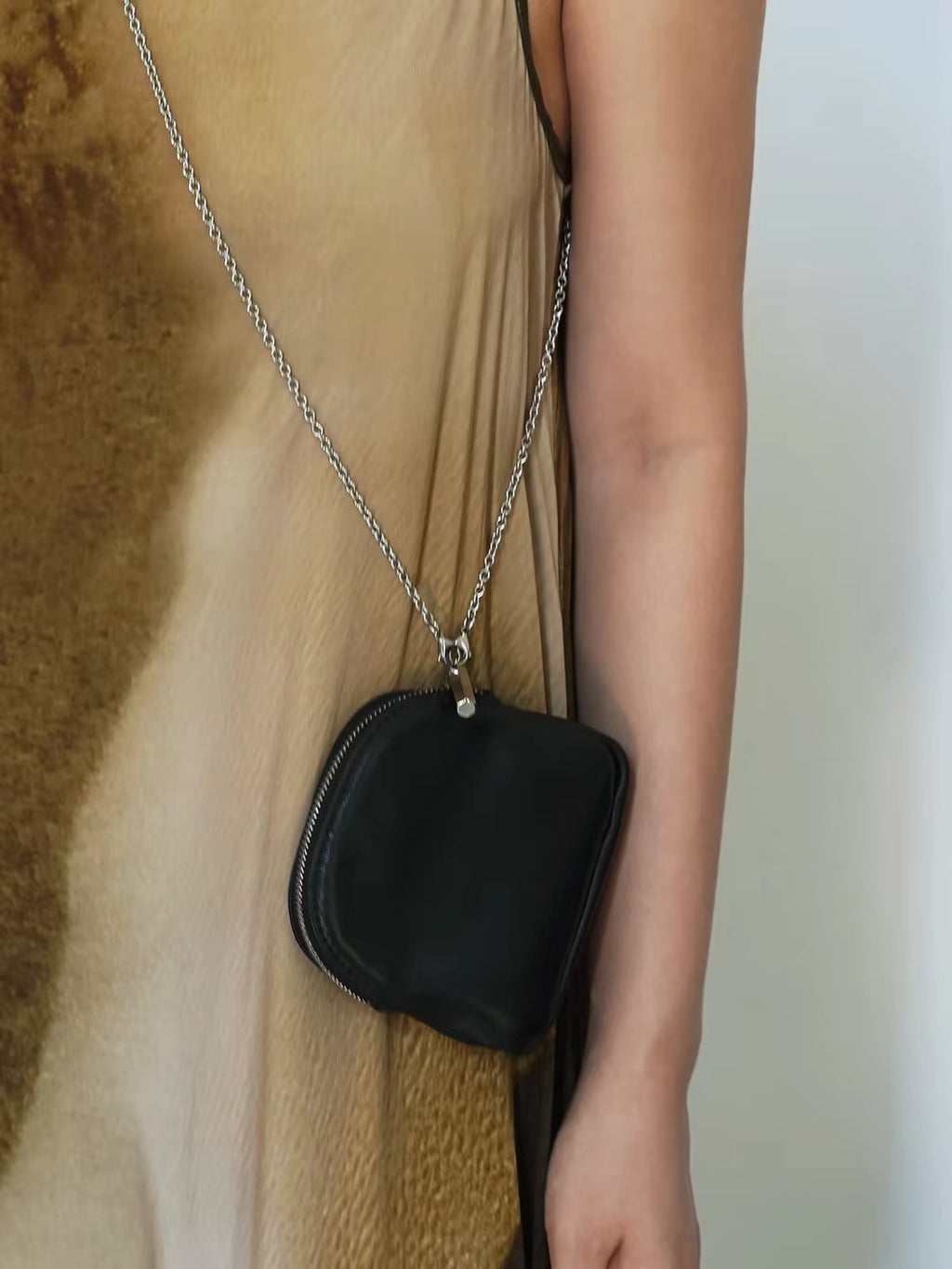 A close-up of a person wearing a black leather crossbody bag with a silver chain strap, styled over a beige and brown patterned dress.