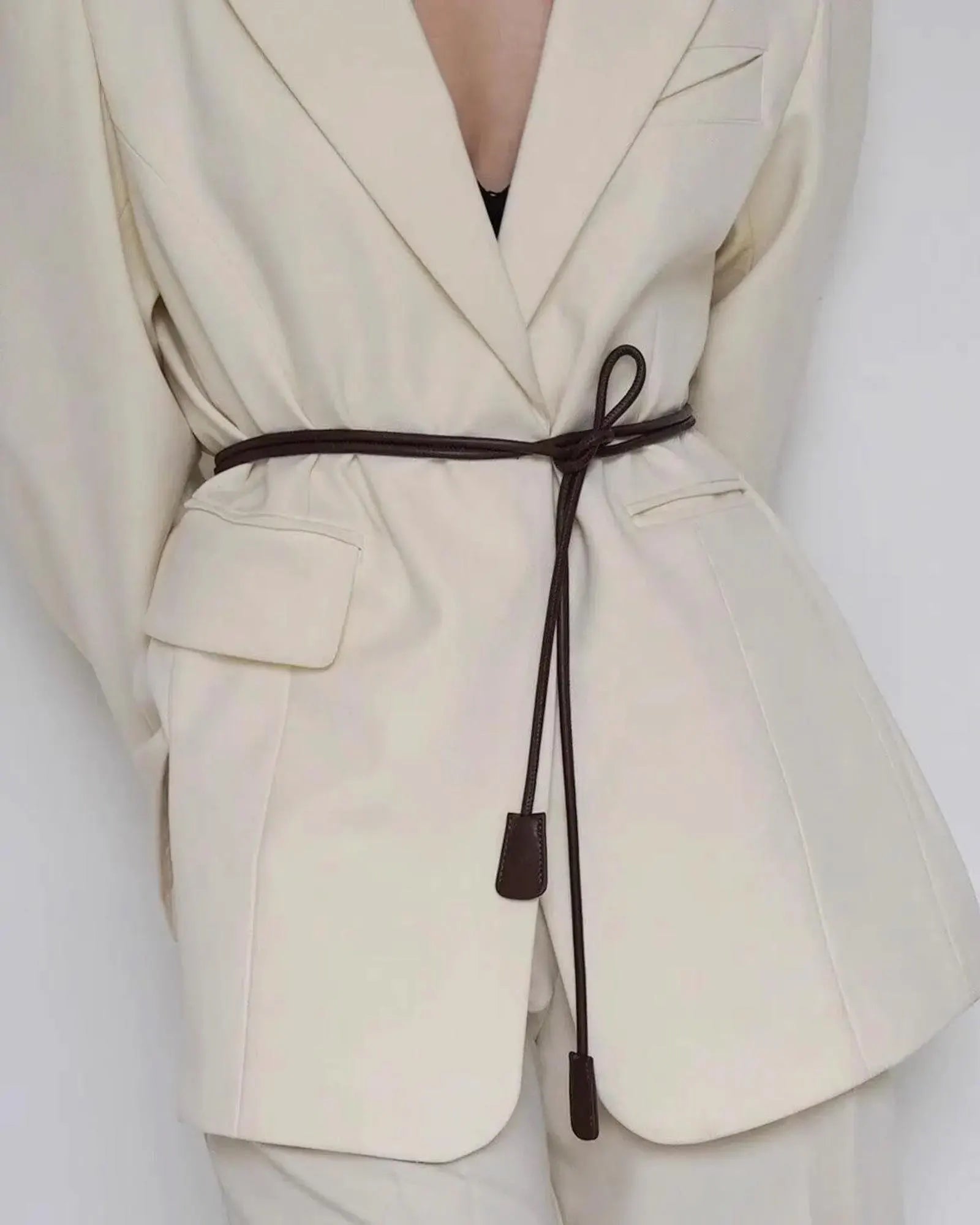 A close-up of a model wearing a cream-colored blazer with a dark brown leather tie belt at the waist, set against a plain light gray background.